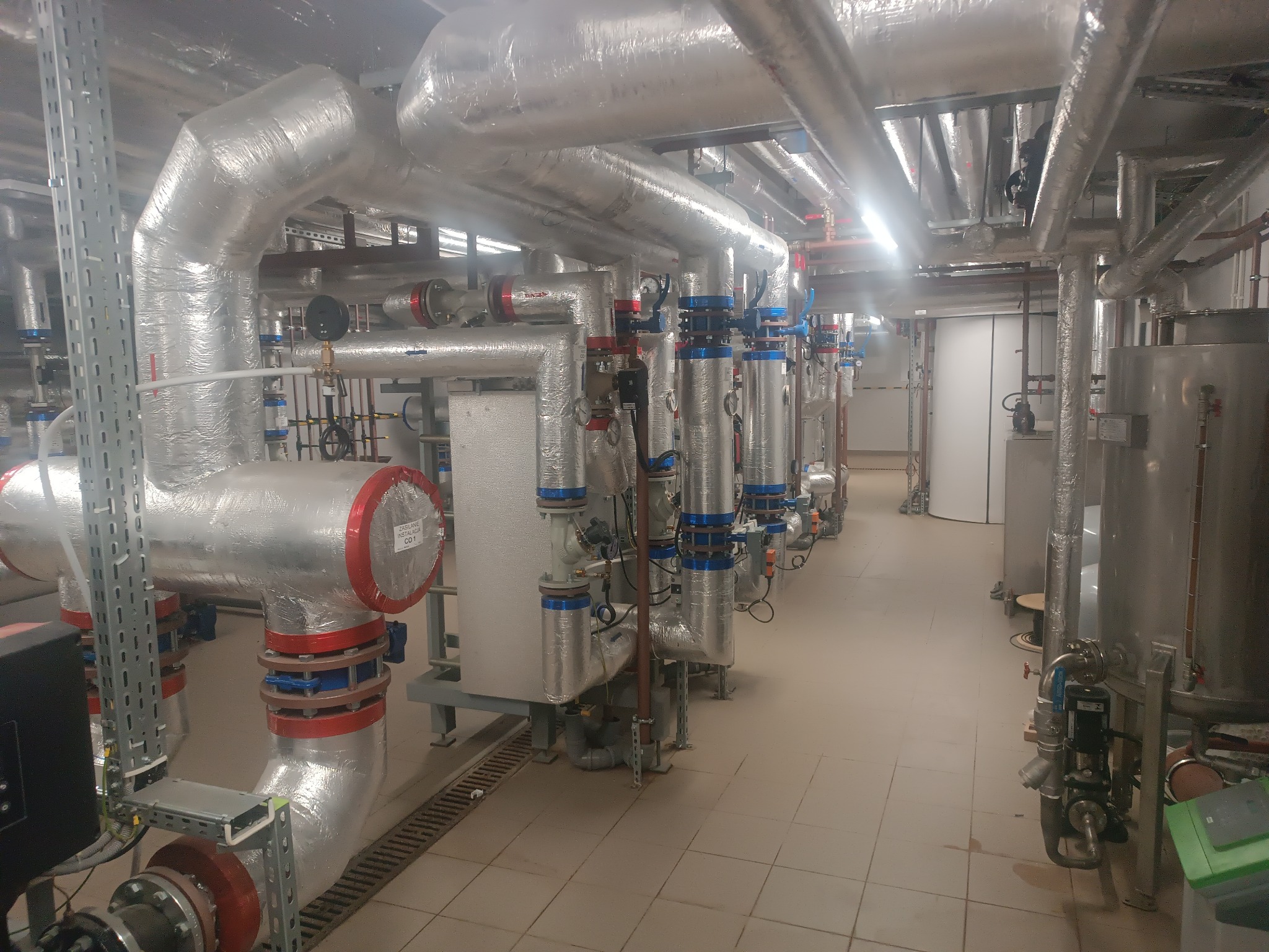 Rzędy izolowanych rur i zaworów w kotłowni, z widocznymi elementami mocującymi i okablowaniem, na tle płytek podłogowych.