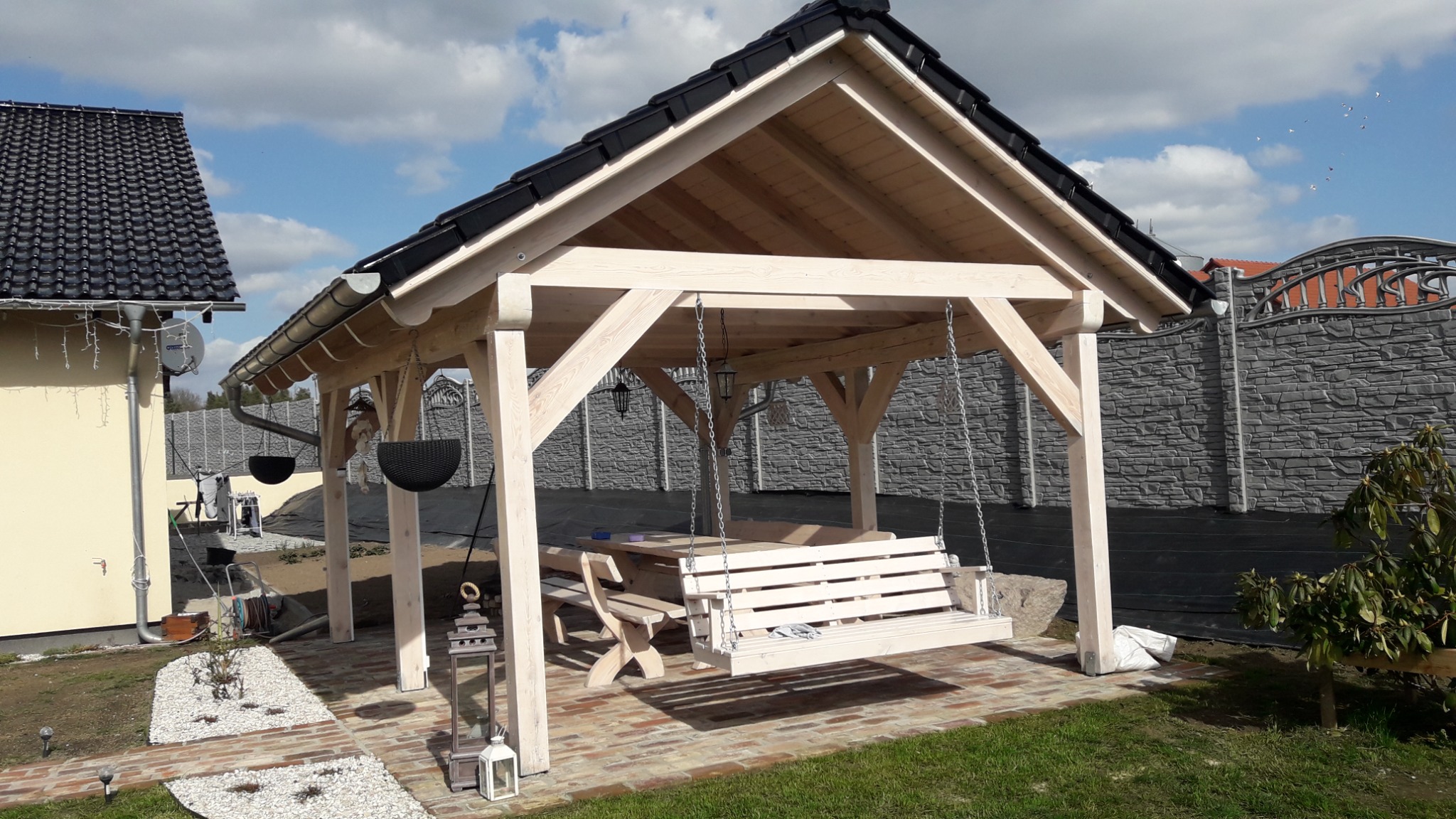 Drewniana altana z huśtawką i stołem, czarny dach, oświetlenie w stylu latarni, otoczenie z kostki brukowej i trawy, widok na dom i ozdobny mur.