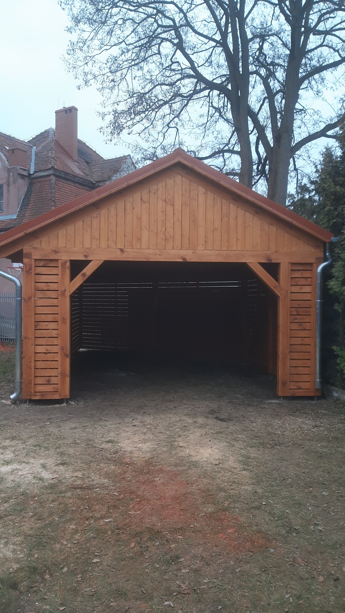 Drewniana wiata garażowa z brązowym dachem i rynnami, widok z zewnątrz na tle trawnika i drzew