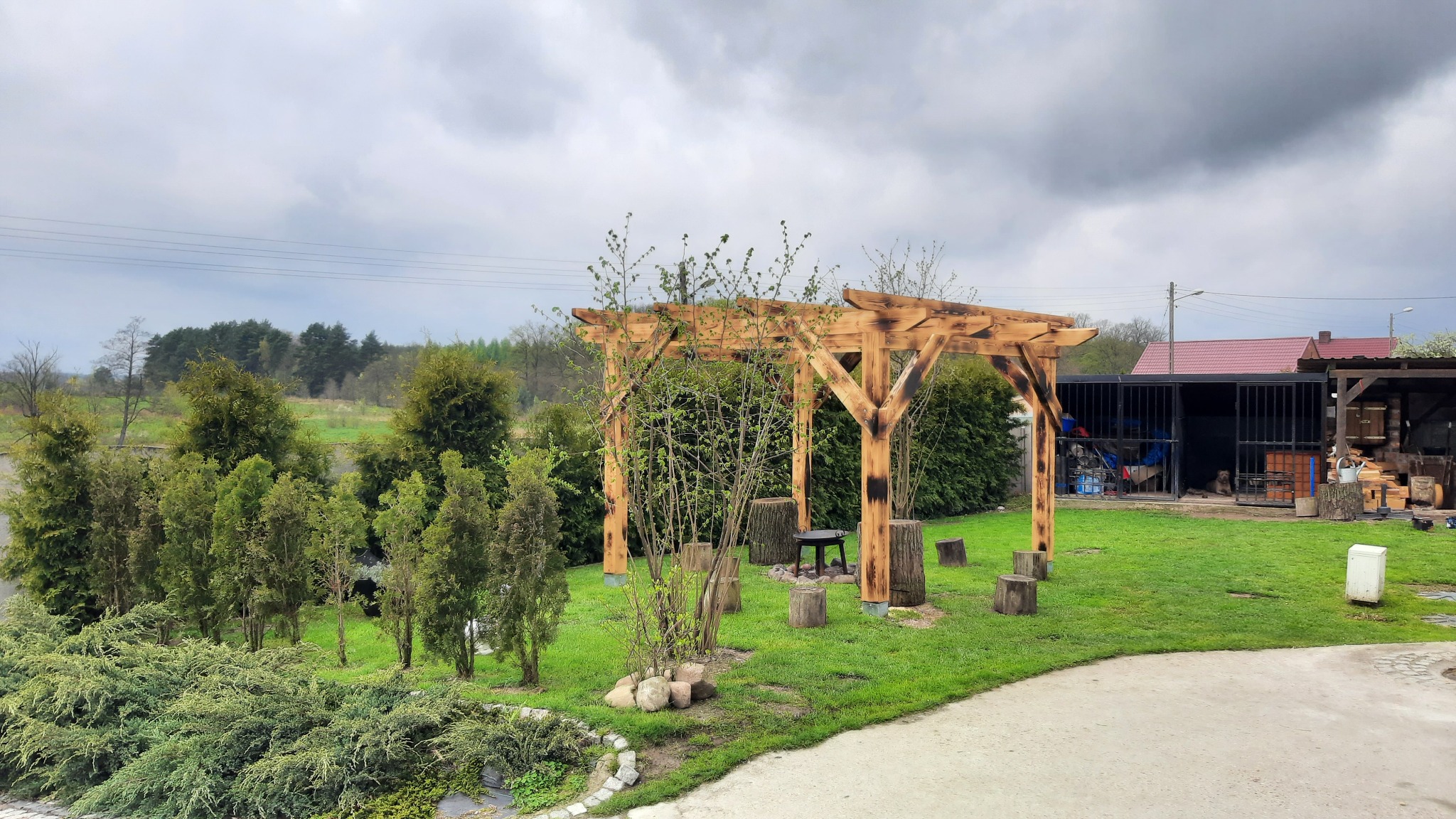 Drewniana pergola ogrodowa z opalonymi słupami, otoczona zielenią i pniakami, na tle pochmurnego nieba i zabudowań gospodarczych.