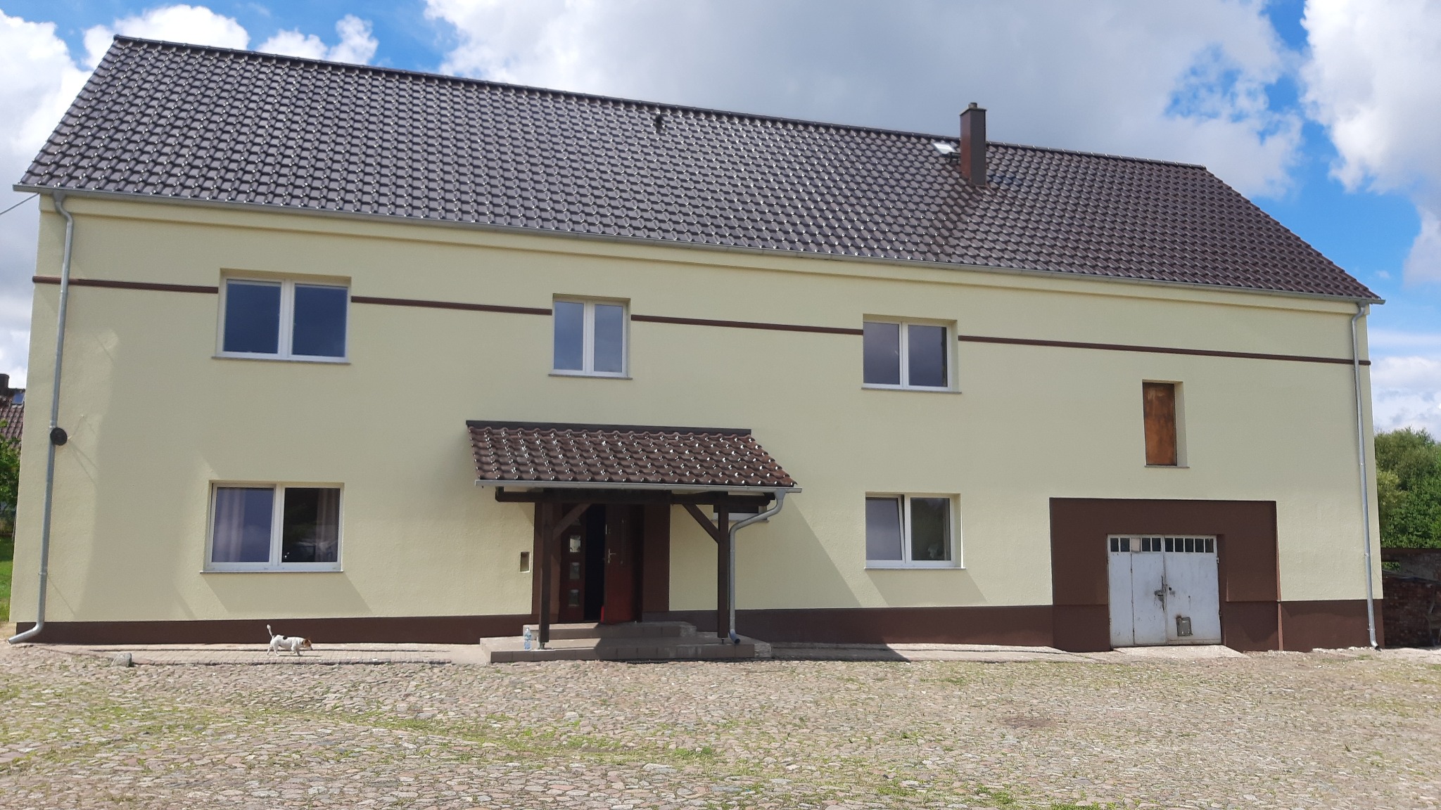 Dwukondygnacyjny budynek w Brzeźnicy z nowym dachem z brązowej dachówki ceramicznej i świeżo pomalowaną elewacją w odcieniach beżu i brązu, widoczny fragment brukowanego podjazdu i mały pies.