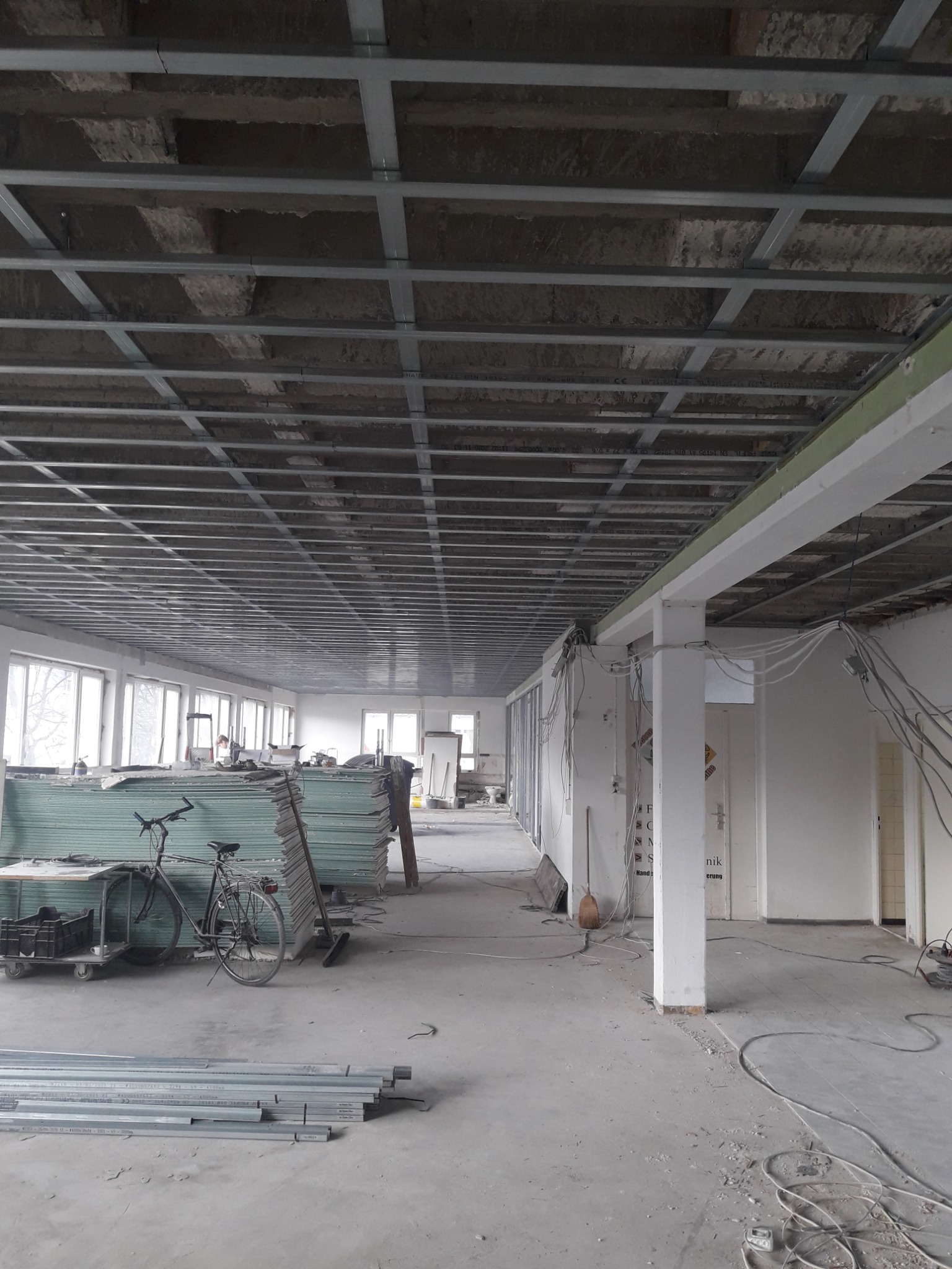 Wnętrze w trakcie prac wykończeniowych: widok na sufit z metalową konstrukcją, stosy płyt gipsowo-kartonowych, rury na podłodze i rower oparty o ścianę.