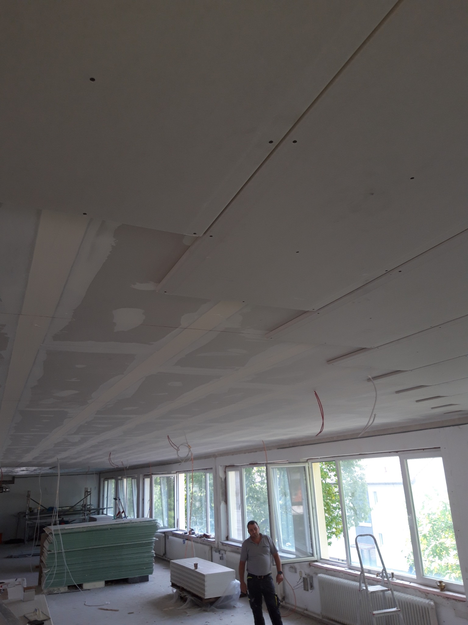 Montaż sufitu podwieszanego z płyt kartonowo-gipsowych w pomieszczeniu z dużymi oknami, widoczne przewody elektryczne i pracownik.