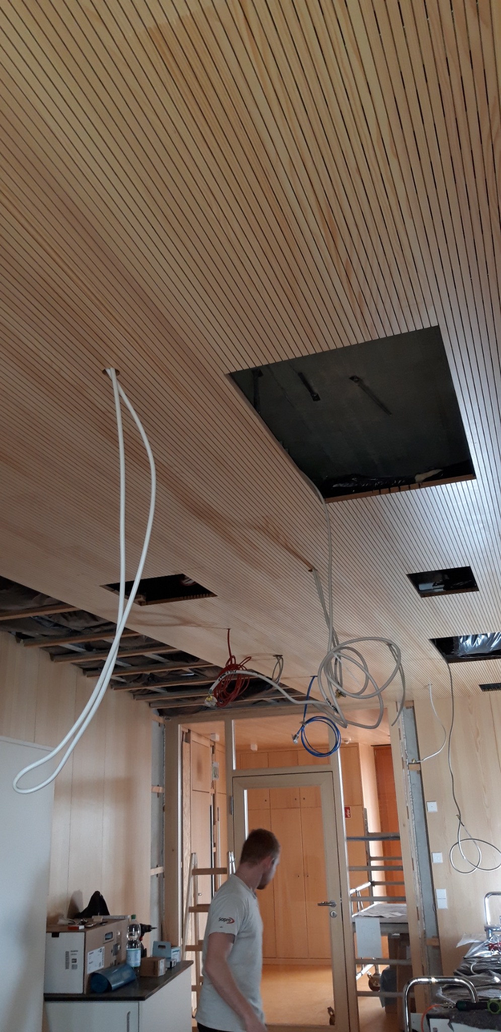 Drewniany sufit listwowy z otworami rewizyjnymi i wystającymi przewodami elektrycznymi, widoczna konstrukcja nośna oraz częściowo widoczna osoba w tle.