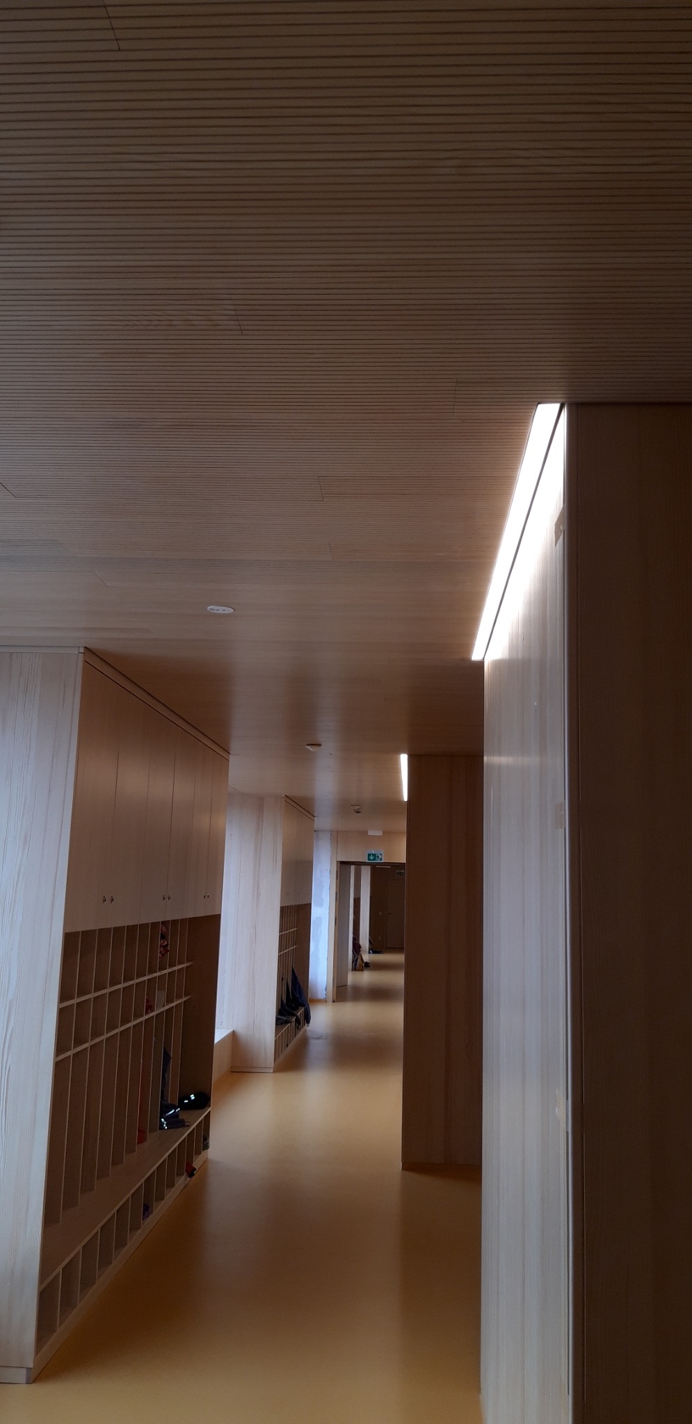 Długi korytarz z drewnianą boazerią na ścianach i suficie, oświetlony liniowymi lampami LED, z szafkami wnękowymi po lewej stronie i żółtą podłogą.