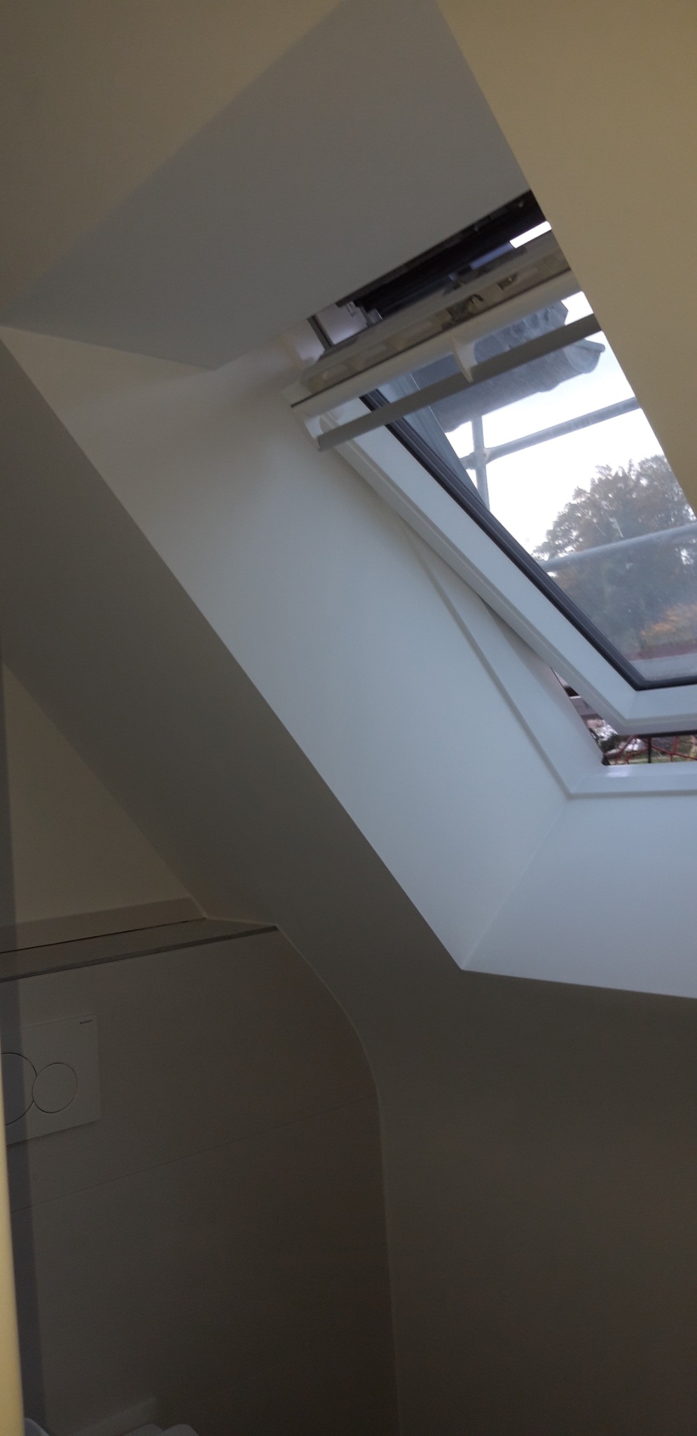 Poddasze z oknem dachowym po malowaniu na biało, widoczne gładkie powierzchnie ścian i skosów, okno uchylone z widokiem na drzewa na zewnątrz.