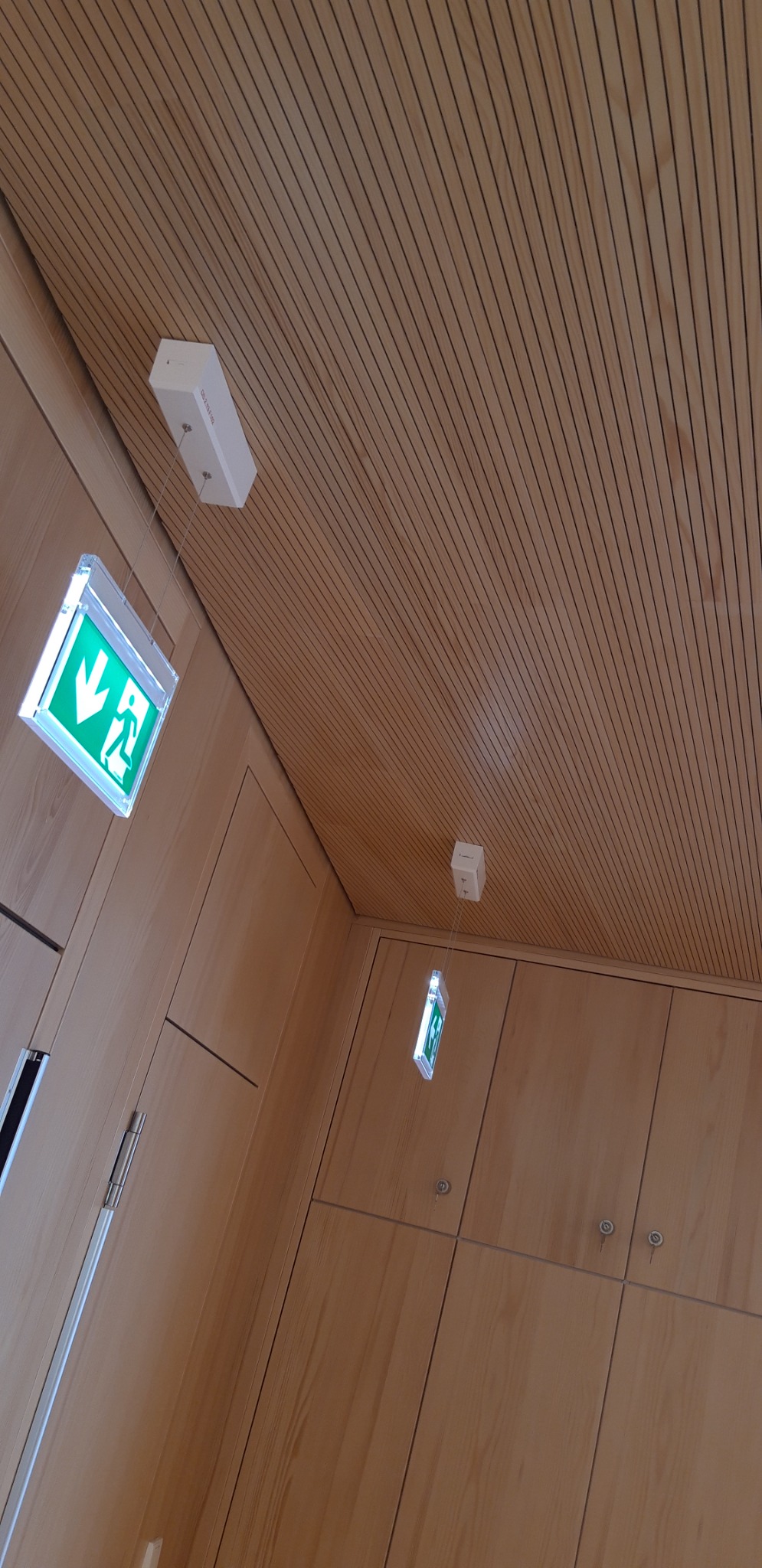 Drewniany sufit lamelowy i ściana z zamontowanymi drzwiami oraz podwieszonymi znakami ewakuacyjnymi z zielonymi strzałkami.