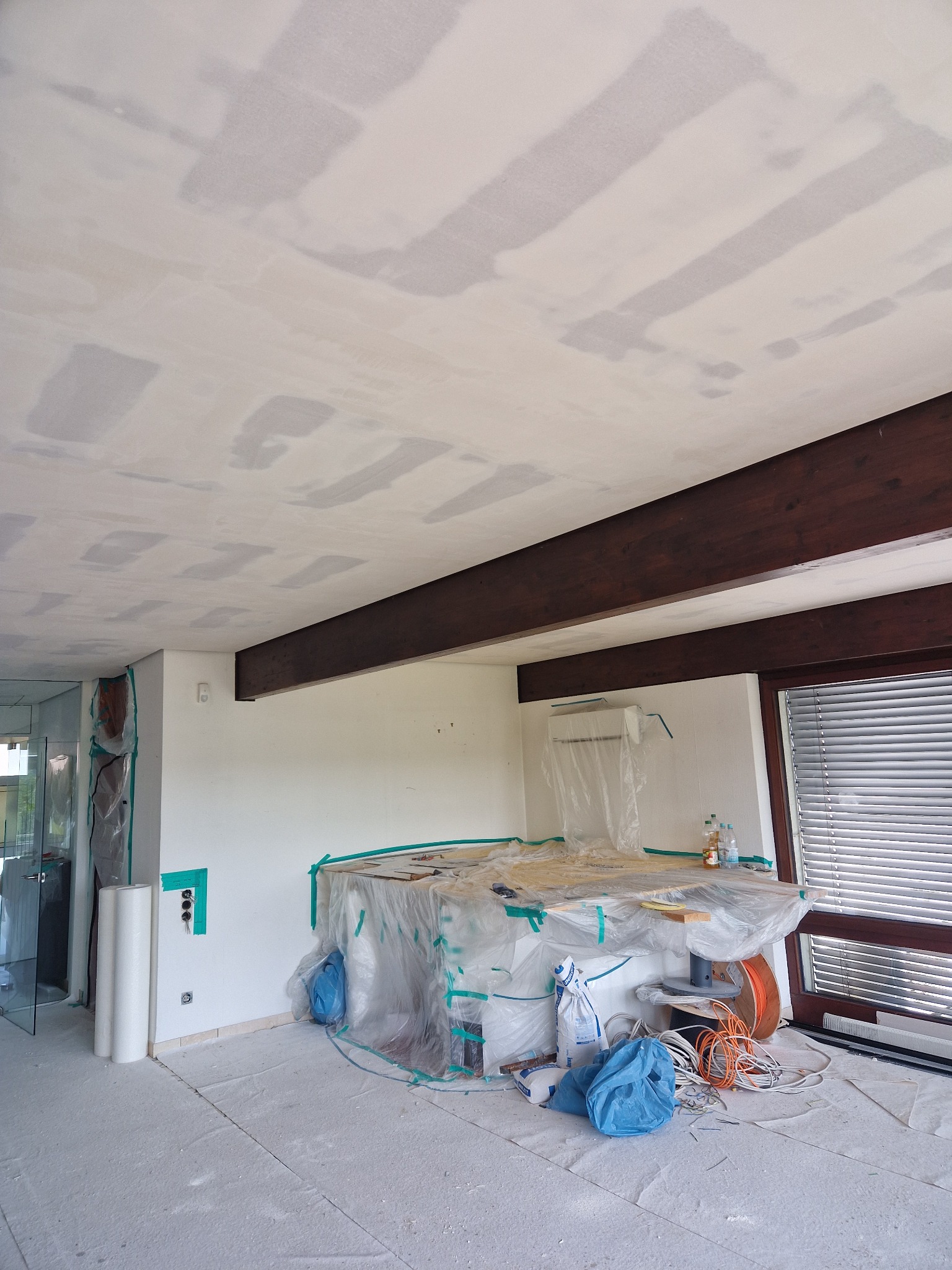 Pokój w trakcie prac wykończeniowych: sufit z widocznymi śladami szpachlowania, zabezpieczone meble folią, podłoga pokryta pyłem, widoczne drewniane belki stropowe i okno z żaluzjami.