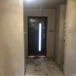 Świeżo nałożone, jasne tynki na ścianach korytarza w trakcie remontu, widoczne puszki elektryczne, zabezpieczone drzwi wejściowe folią ochronną i surowa podłoga.