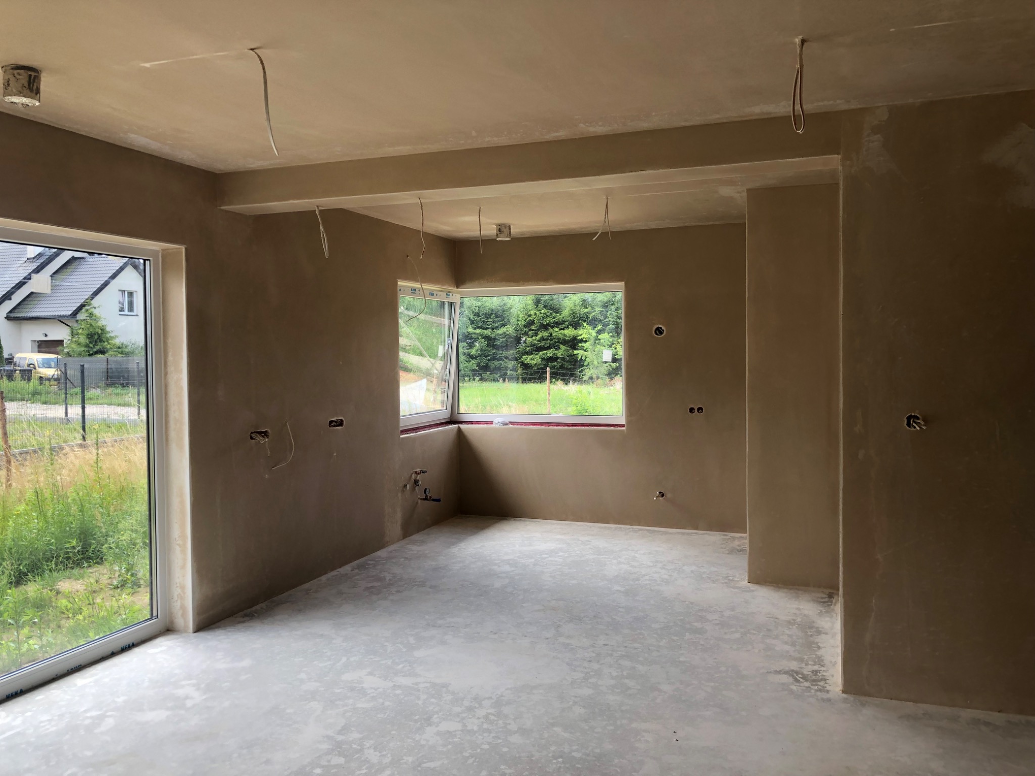 Wnętrze surowego pomieszczenia po nałożeniu tynków, widoczne wystające kable, okna z widokiem na zieleń, ściany w kolorze beżowym, surowy beton na podłodze.