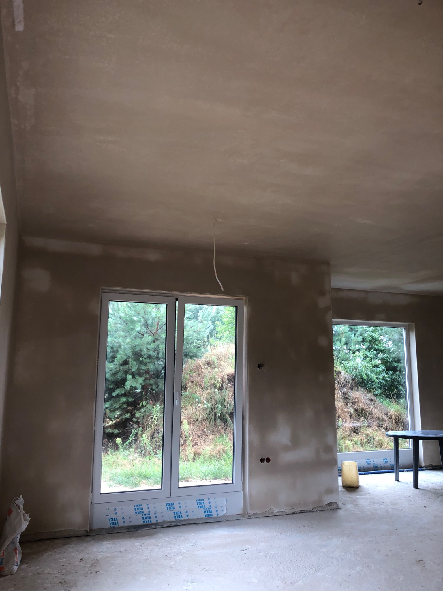 Wnętrze w trakcie prac tynkarskich: ściany i sufit pokryte świeżym, jasnym tynkiem, widoczne okna z widokiem na zieleń, na podłodze resztki materiałów budowlanych.