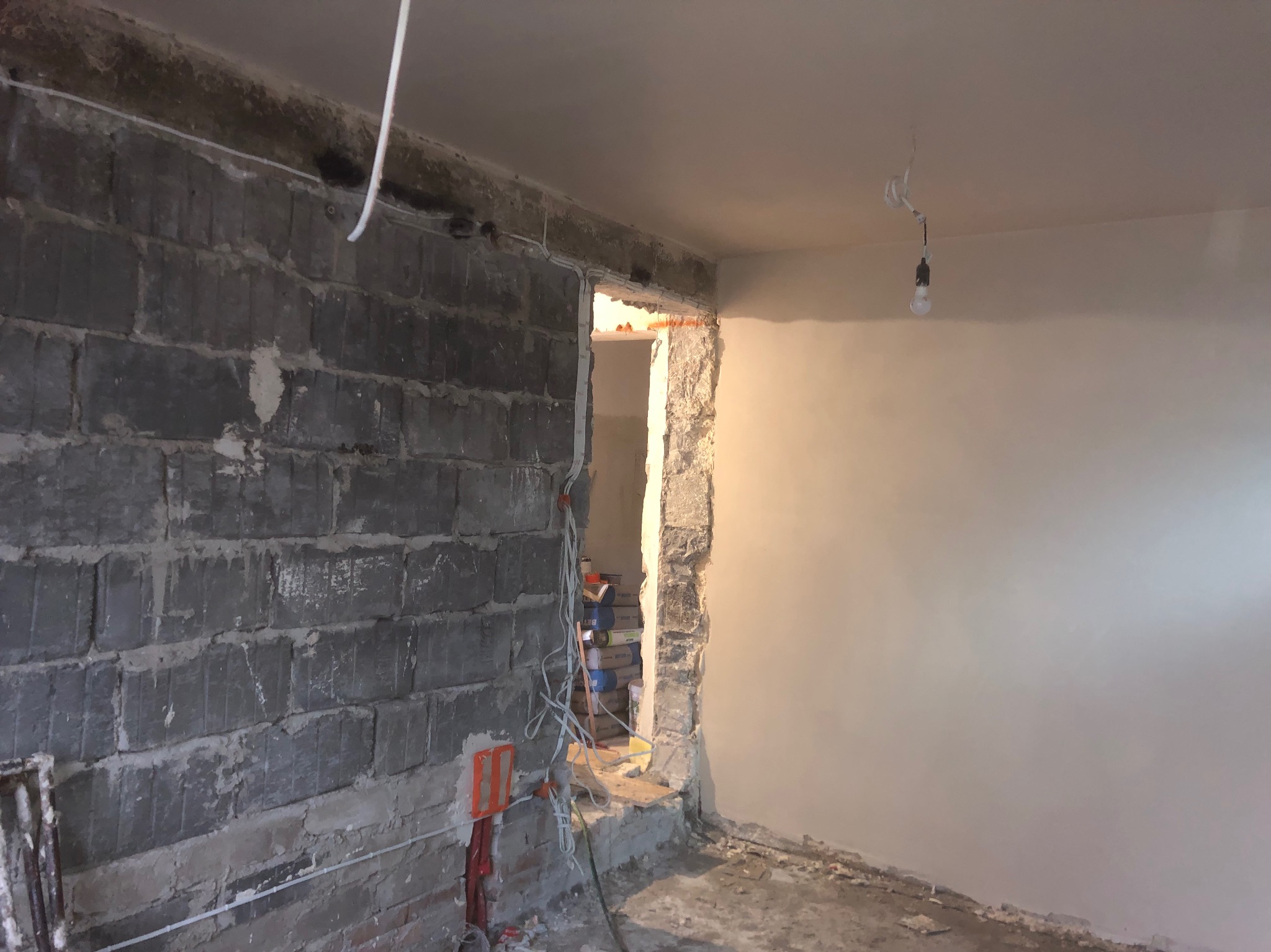 Wnętrze w trakcie remontu: surowa ściana z bloczków obok fragmentu ściany po nałożeniu gładzi, widoczne przewody elektryczne i worki z zaprawą w przejściu.