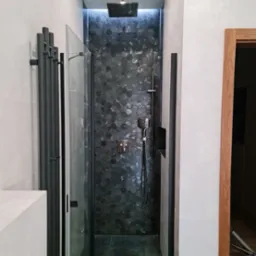 Nowoczesna kabina prysznicowa z heksagonalną mozaiką w odcieniach szarości i czerni, deszczownicą sufitową i panelem natryskowym, uzupełniona grzejnikiem drabinkowym w kolorze antracytowym.