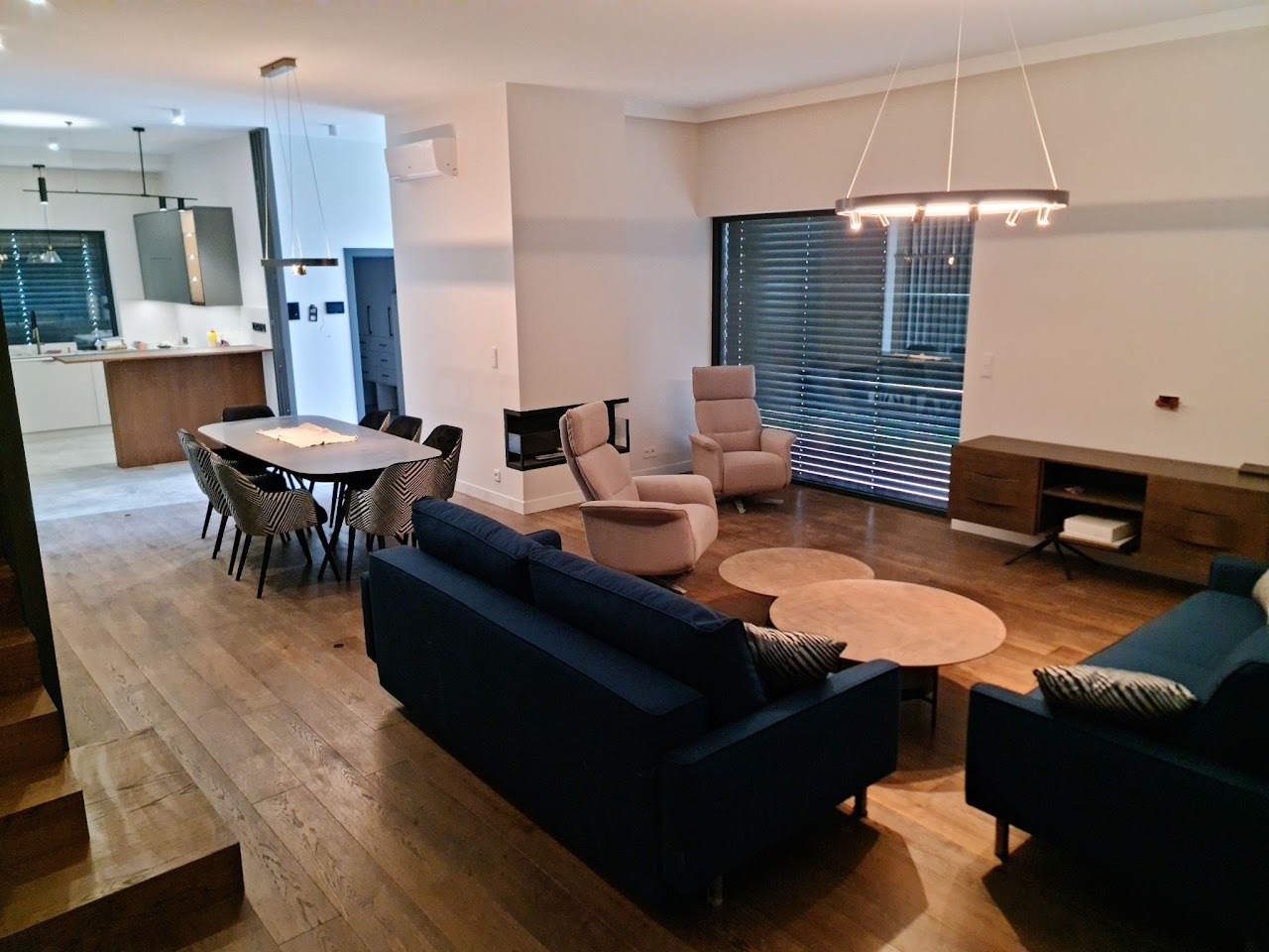 Przestronne wnętrze domu z drewnianą podłogą, obejmujące salon z ciemnoniebieskimi sofami, okrągłymi stolikami kawowymi, beżowymi fotelami, szafką RTV, jadalnię ze stołem i krzesłami w geometryczne...