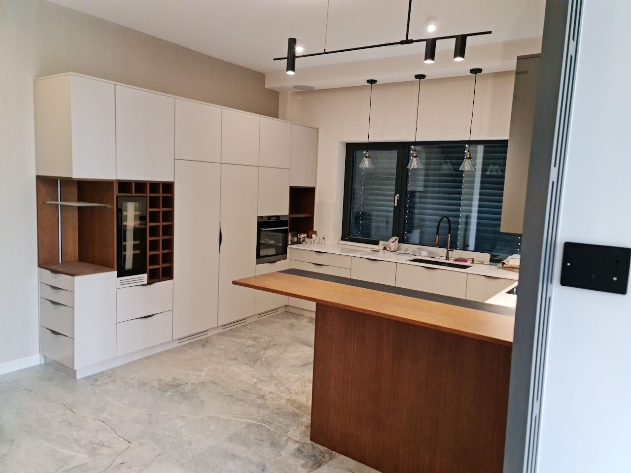 Nowoczesna kuchnia z białymi szafkami, drewnianym barkiem i marmurową podłogą; oświetlenie punktowe i wiszące lampy nad oknem.