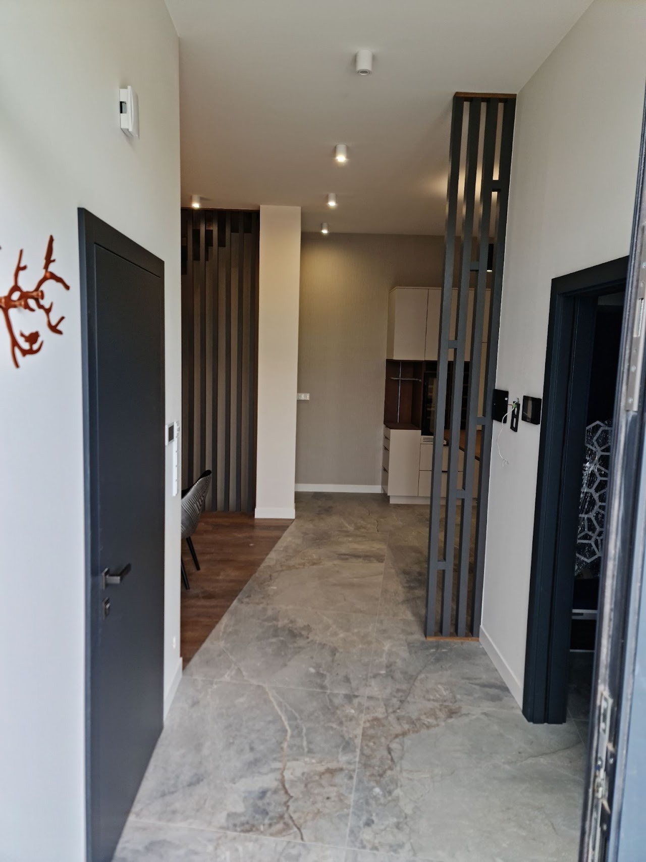 Nowoczesny przedpokój z szarą podłogą imitującą marmur, ciemnoszare drzwi i dekoracyjna ścianka działowa z pionowych listew, widok na wnętrze mieszkania z oświetleniem punktowym.