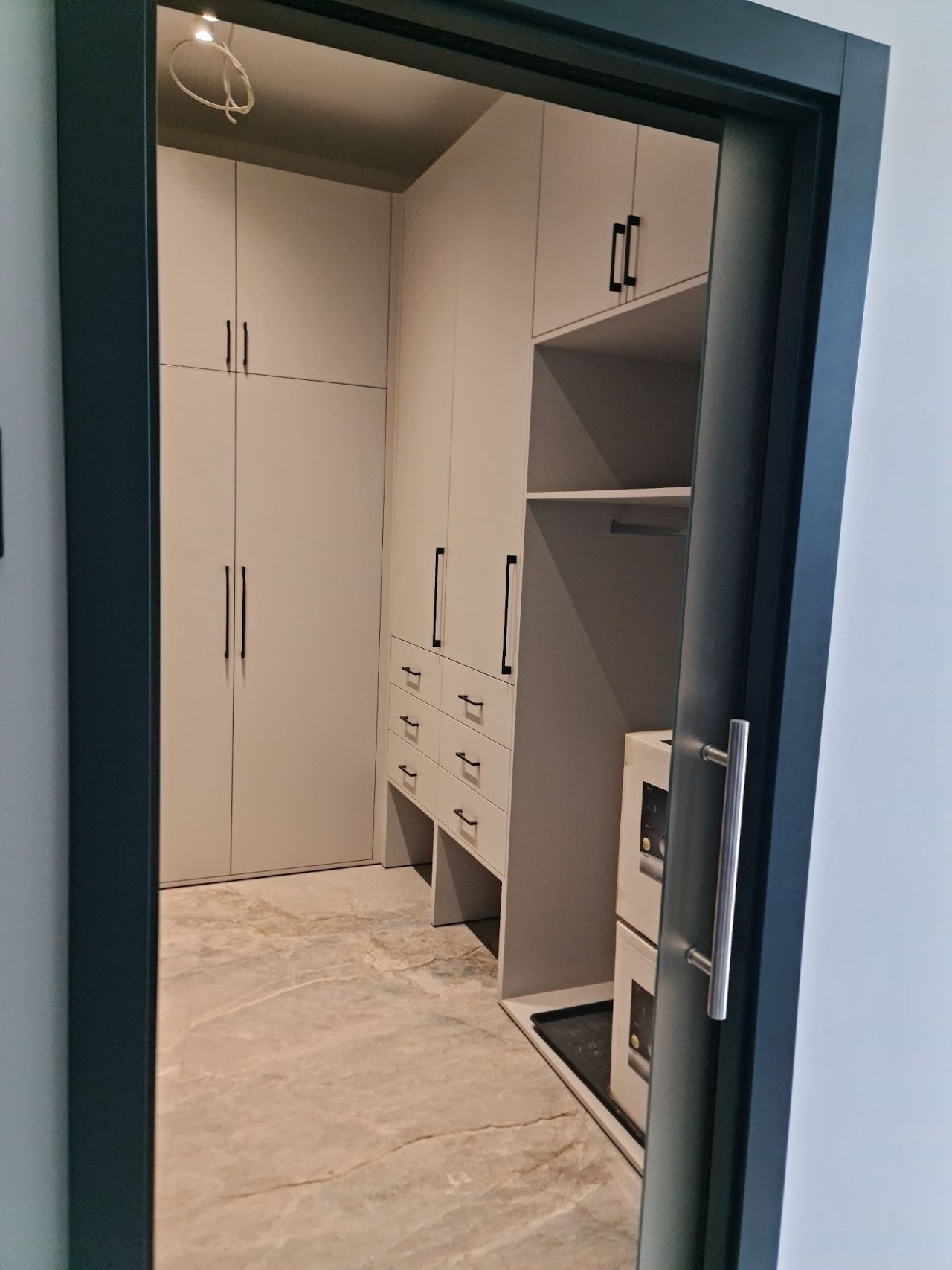 Widok na wnętrze garderoby z szarymi szafami z czarnymi uchwytami i marmurową podłogą, widziane przez otwarte, grafitowe drzwi z srebrną klamką.