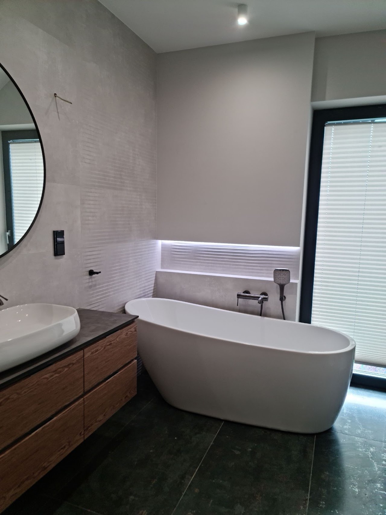 Nowoczesna łazienka z wolnostojącą wanną, umywalką nablatową na drewnianej szafce i szarą płytką imitującą beton z subtelnym wzorem, oświetlona listwą LED nad wanną.