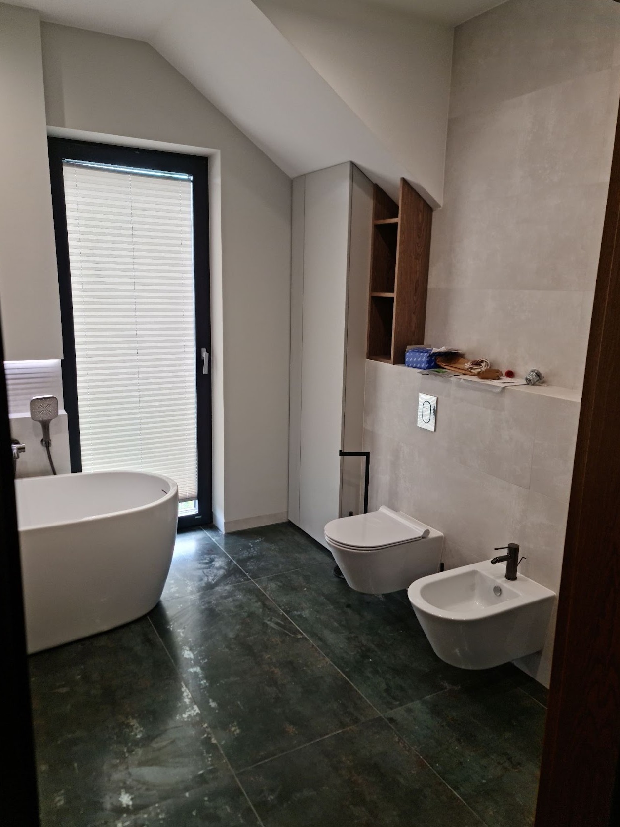 Nowoczesna łazienka z wanną wolnostojącą, toaletą i bidetem na tle ściany z szarych płytek i ciemnej podłogi. W tle okno z żaluzjami plisowanymi i szafka z drewnianymi półkami.