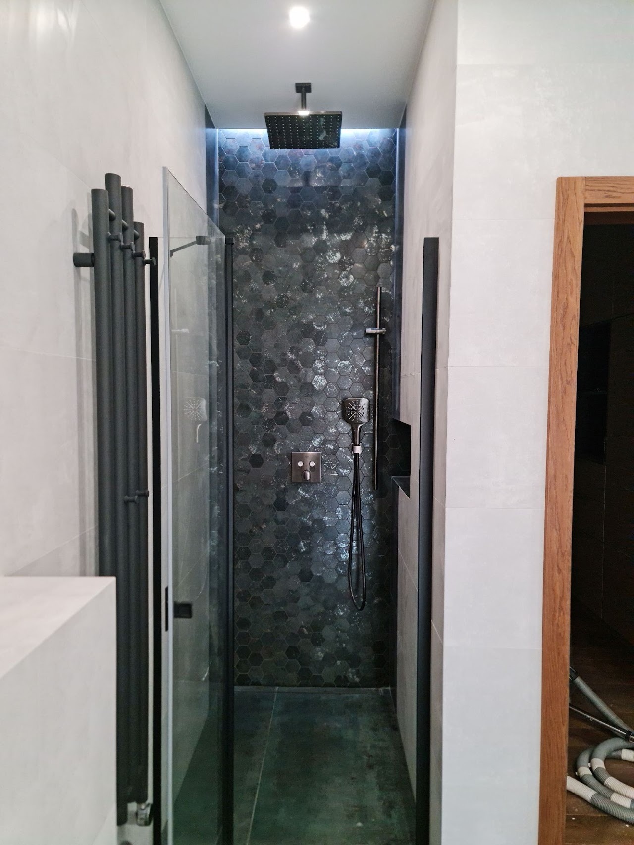 Nowoczesna kabina prysznicowa z heksagonalną mozaiką w odcieniach szarości i czerni, deszczownicą sufitową i panelem natryskowym, uzupełniona grzejnikiem drabinkowym w kolorze antracytowym.