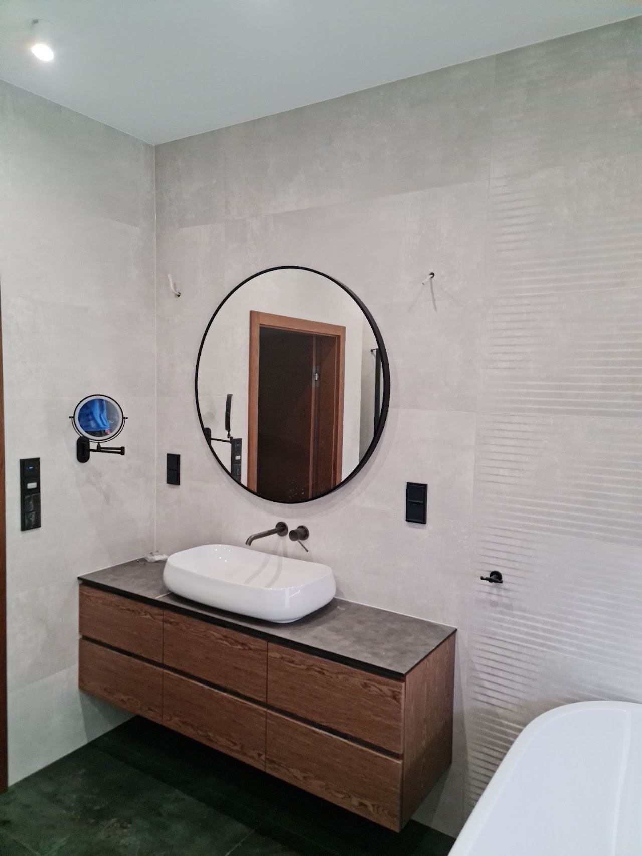 Nowoczesna łazienka z umywalką nablatową, okrągłym lustrem w czarnej ramie, drewnianą szafką pod umywalką i szarą płytką imitującą beton na ścianach, z widocznym fragmentem wanny.