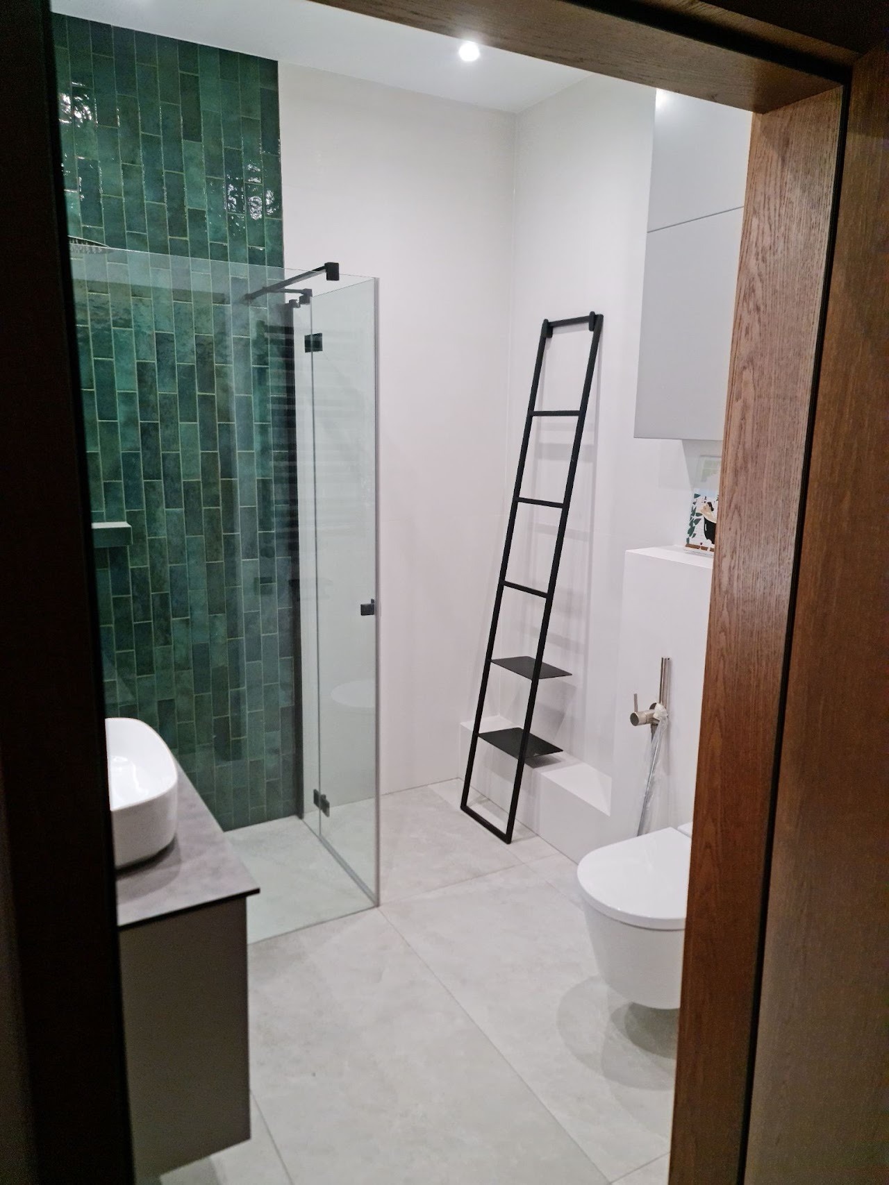 Nowoczesna łazienka z kabiną prysznicową wyłożoną zielonymi, podłużnymi płytkami, białą umywalką nablatową na szarej szafce, wiszącą toaletą i czarnym, metalowym regałem-drabinką.