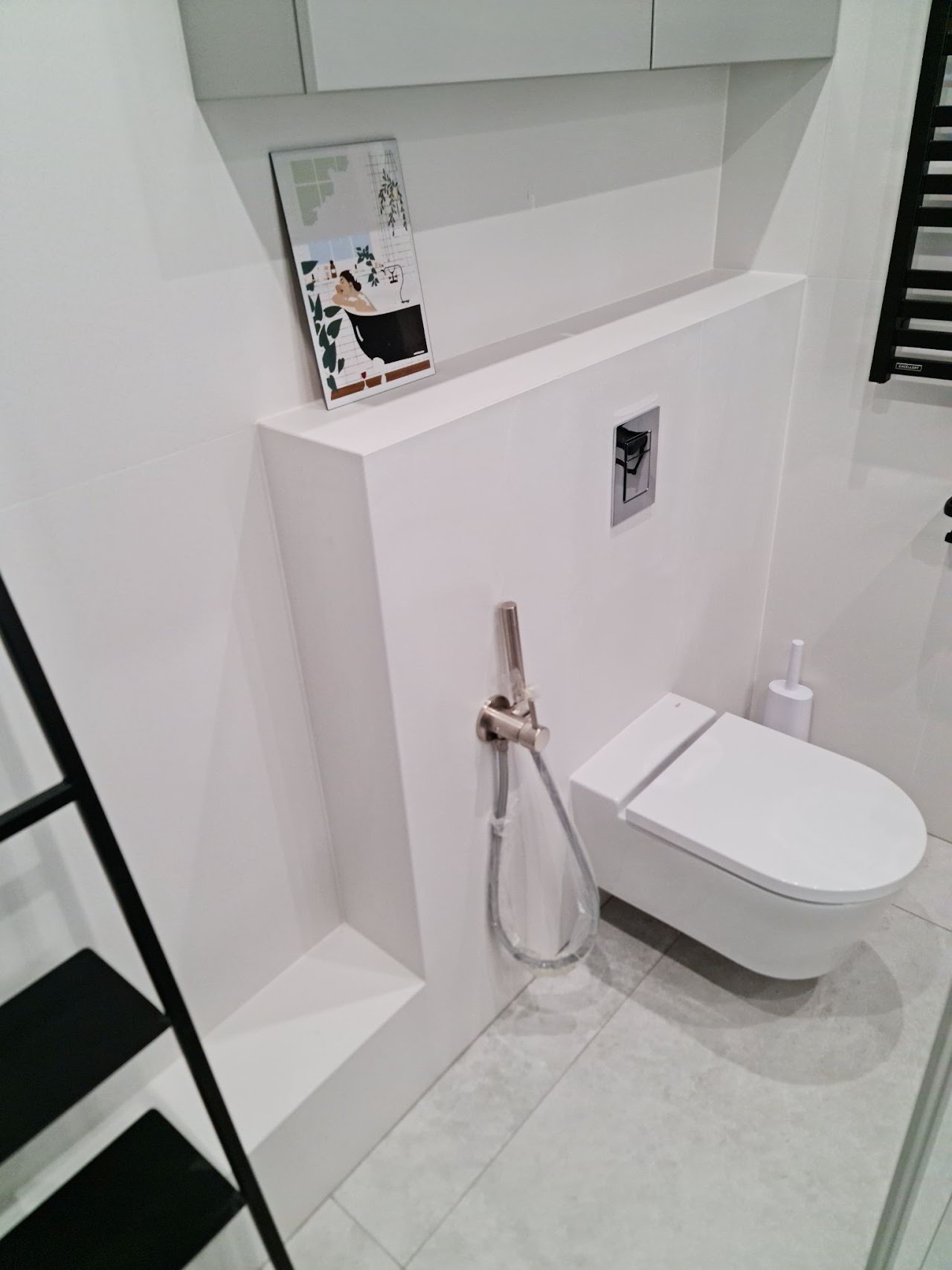 Nowoczesna, minimalistyczna łazienka z podwieszaną toaletą, bidetką w kolorze szczotkowanego niklu, i dekoracyjnym obrazkiem z motywem kobiety w wannie, umieszczonym na półce z zabudowy...