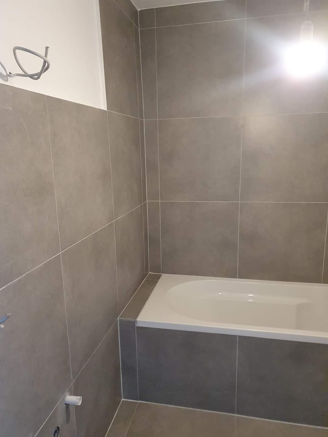 Szare płytki ceramiczne ułożone na ścianach i obudowie wanny w łazience w trakcie wykańczania, widoczne wystające kable i rurki.