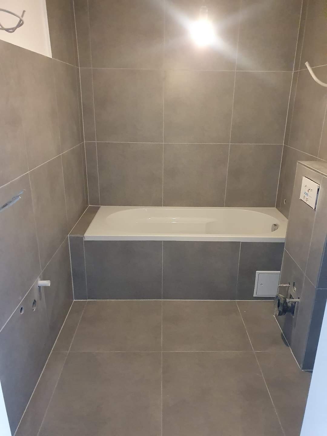 Wykończona łazienka z szarymi płytkami, białą wanną i widocznymi przyłączami wodno-kanalizacyjnymi przed montażem armatury.