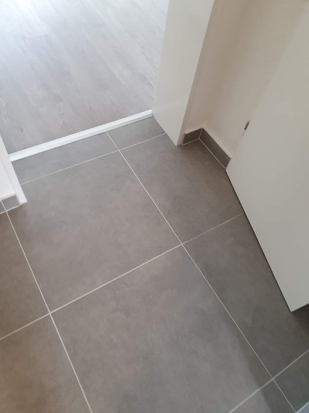 Połączenie podłogi z szarych płytek ceramicznych z jasnymi panelami podłogowymi, widoczna listwa przypodłogowa i fragment drzwi.