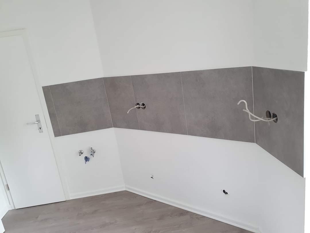 Fragment wnętrza z szarymi płytkami na ścianie, wystającymi przewodami elektrycznymi i widoczną białą ścianą oraz fragmentem podłogi wyłożonej panelami podłogowymi.