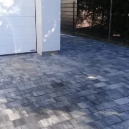 Ułożona szara kostka brukowa przed białym garażem z bramą segmentową, widoczny fragment ogrodzenia i roślinność w tle.