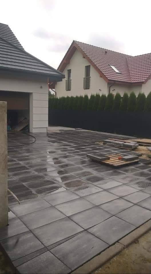 Świeżo ułożona, mokra, szara kostka brukowa na podjeździe, z paletą z kostką w tle, widoczny dom z garażem i iglaki wzdłuż ogrodzenia.