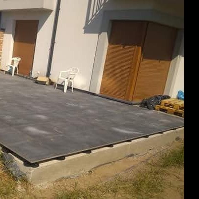 Przygotowanie podłoża pod taras z płyt betonowych na podwyższeniu, widoczne regulowane wsporniki, w tle jasny budynek z brązowymi drzwiami i dwoma białymi krzesłami.