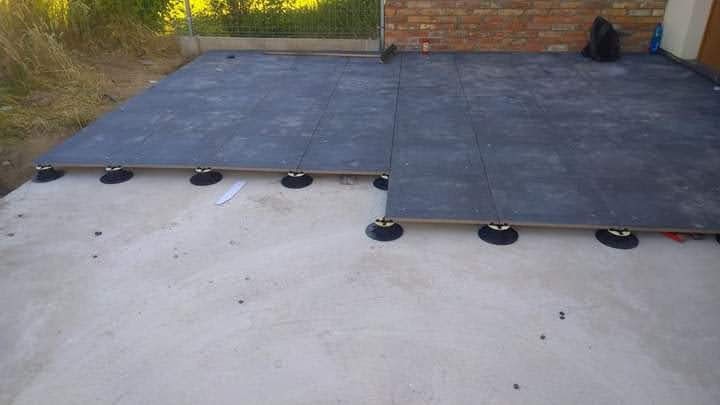 Montaż podłogi tarasowej z płyt na regulowanych wspornikach. Widoczne płyty ułożone na betonowej wylewce, część płyt jeszcze nie zamontowana. W tle mur z cegły.