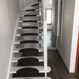 Białe, kręcone schody z czarnymi nakładkami antypoślizgowymi na stopniach, widoczne z dołu, z fragmentem zabudowy szafy w tle.