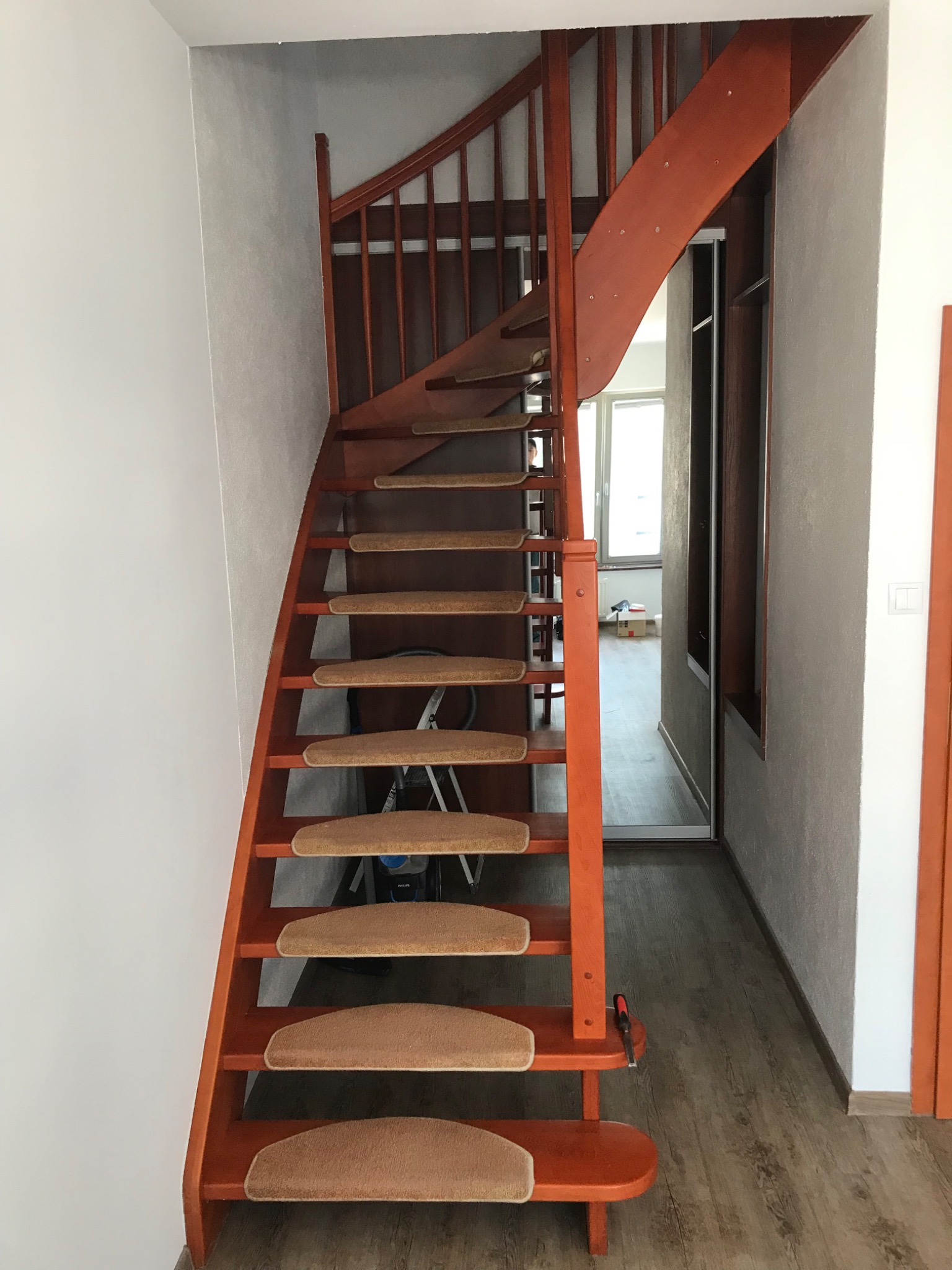 Drewniane schody o pomarańczowym odcieniu z dywanowymi nakładkami na stopniach, widoczne z dołu w wąskim korytarzu, z narzędziem (wkrętakiem) leżącym na jednym ze stopni.