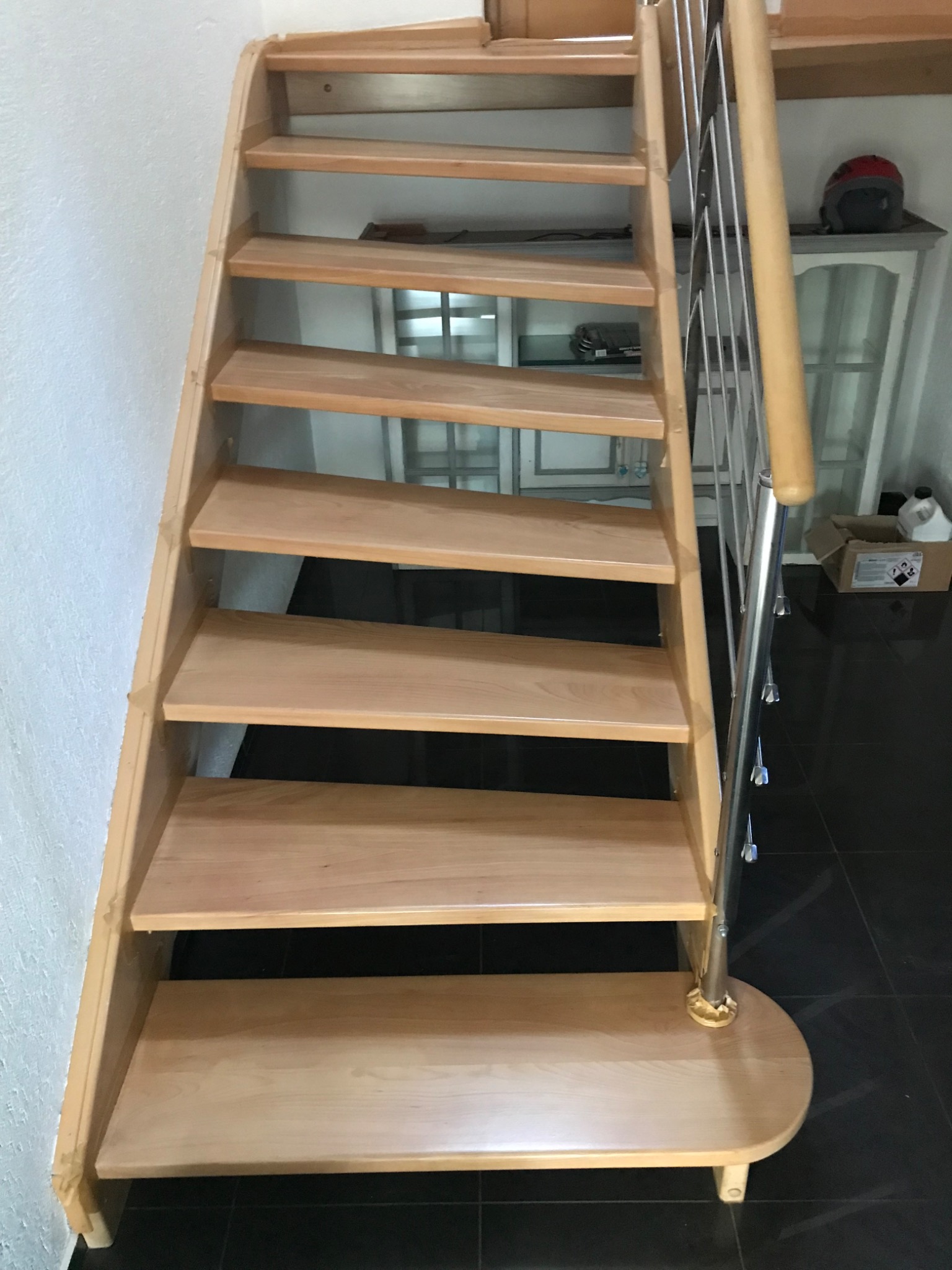 Drewniane schody z metalową balustradą, jasne drewno, widok z dołu, czarne płytki podłogowe, minimalistyczny design, w tle szafka z przeszklonymi drzwiami.