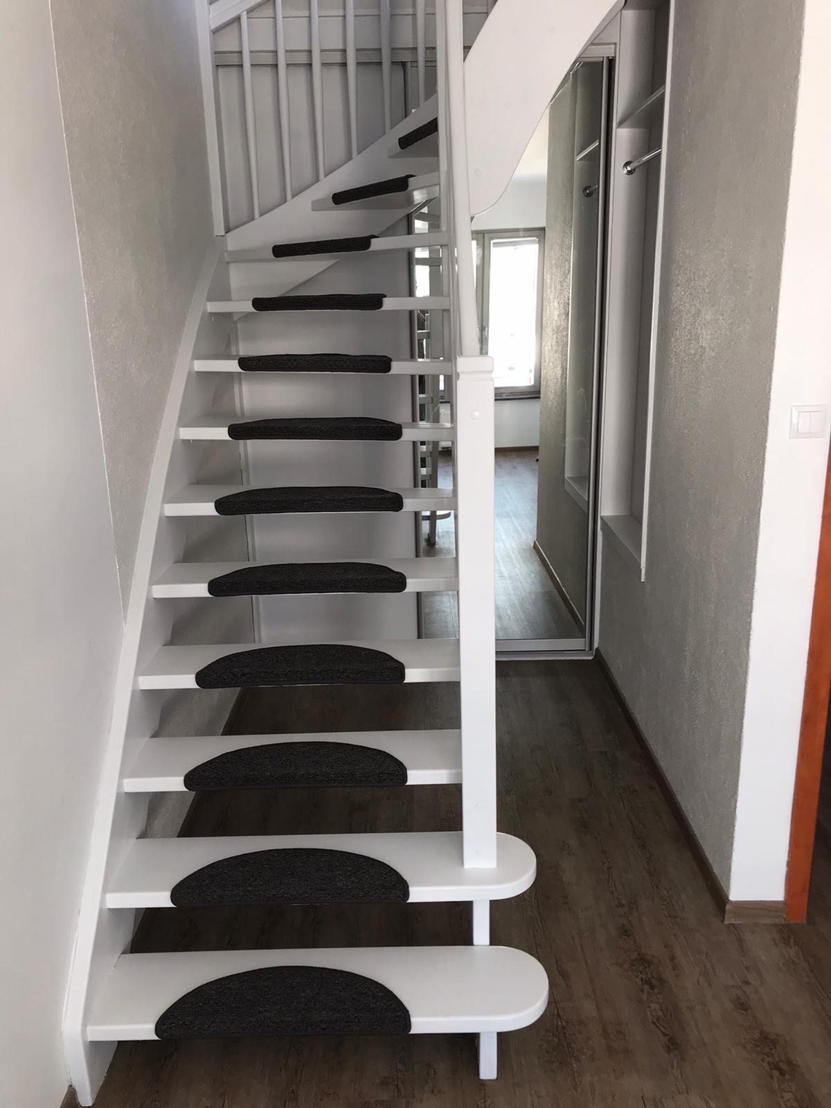 Białe, kręcone schody z czarnymi nakładkami antypoślizgowymi na stopniach, widoczne z dołu, z fragmentem zabudowy szafy w tle.