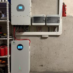 ARD-SUN Sławomir Jackowski - Biały inwerter i magazyn energii Ecoluna na tle surowej ściany. Widoczne zabezpieczenia elektryczne i fragment regału. Instalacja w pomieszczeniu technicznym.