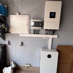 ARD-SUN Sławomir Jackowski - Biały inwerter i magazyn energii Deye zamontowane na szarej ścianie w garażu. Widoczne kable i przewody. Na ścianie skrzynka elektryczna i gniazda.