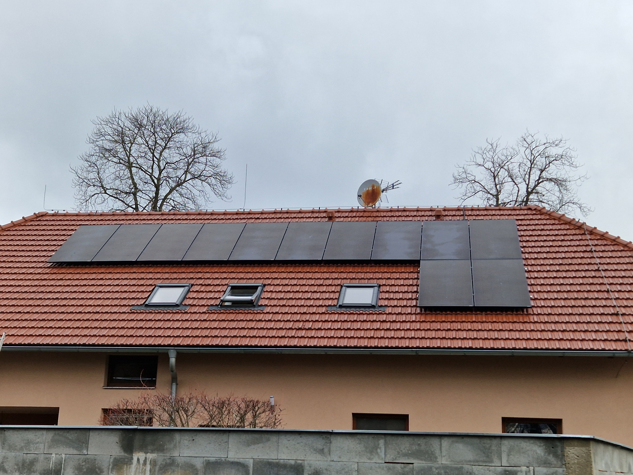 Czerwony dach z dachówkami i oknami dachowymi, na którym zainstalowano panele słoneczne. Na dachu widoczna antena satelitarna, a w tle drzewa bez liści i pochmurne niebo.