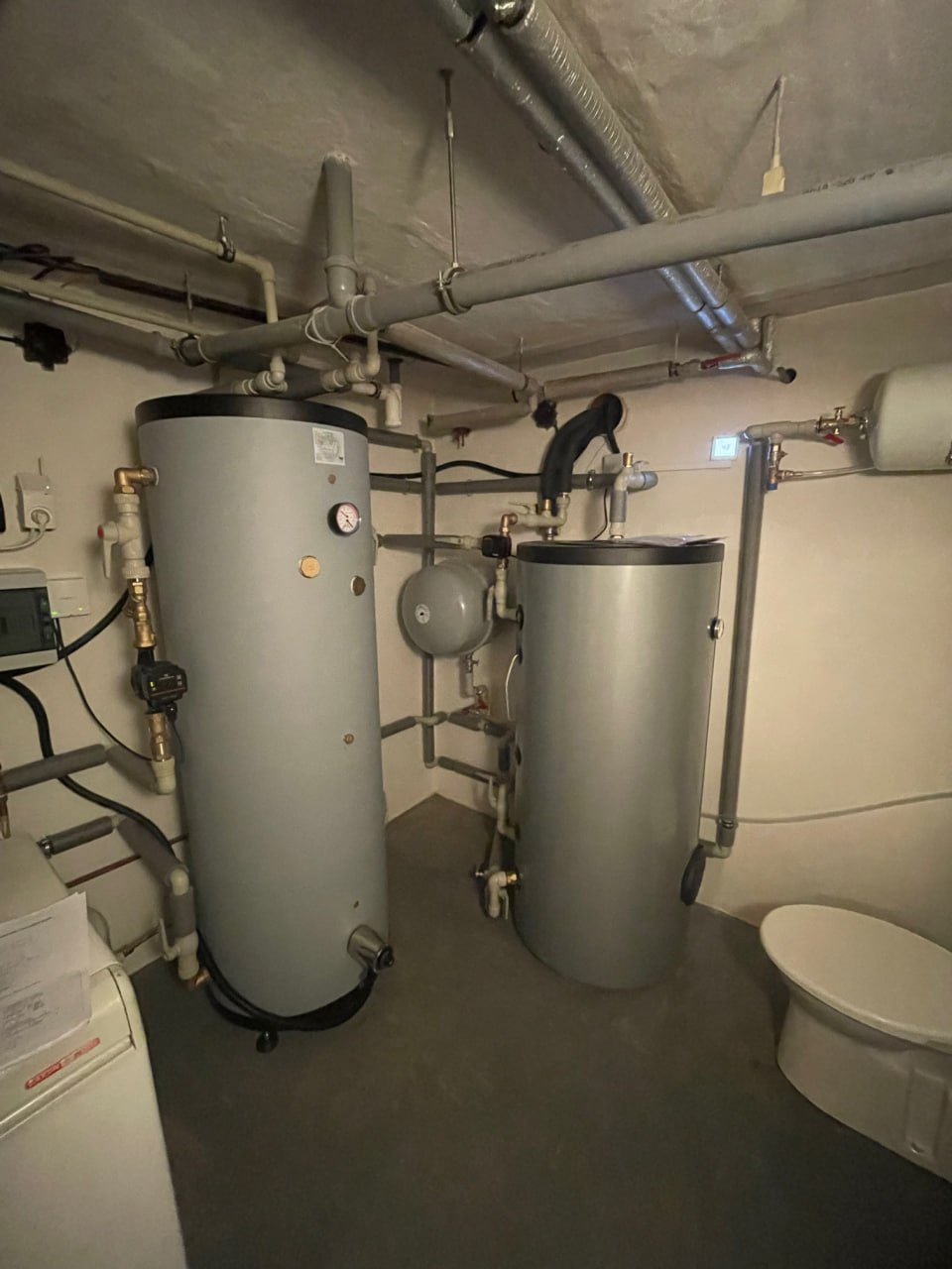 Dwa szare zasobniki ciepłej wody w piwnicy, otoczone rurami, z widocznym manometrem na jednym z nich.