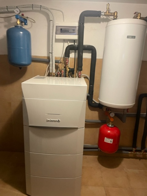 Kompaktowy, kondensacyjny kocioł grzewczy De Dietrich z podłączonym zasobnikiem ciepłej wody i naczyniami wzbiorczymi w kotłowni z beżowymi płytkami na ścianach i podłodze.