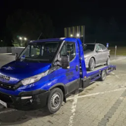 Niebieska laweta Iveco z załadowanym srebrnym autem na parkingu nocą, widoczne logo firmy i oświetlenie parkingu.