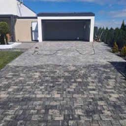 Czyszczenie kostki brukowej myjką ciśnieniową na podjeździe przed garażem. Widoczny efekt czyszczenia, ciemniejsza i jaśniejsza kostka. Kabel myjki na kostce.