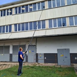 RT Steam - Mężczyzna myje okna na wysokości za pomocą długiej lancy. Budynek z szarymi drzwiami i zielonym trawnikiem w tle. Błękitne niebo.