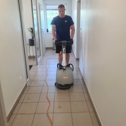 RT Steam - Pracownik z maszyną do czyszczenia podłóg w korytarzu biurowym. Widoczne płytki podłogowe i białe ściany. Pomarańczowy kabel maszyny. Ujęcie z przodu, na wprost.