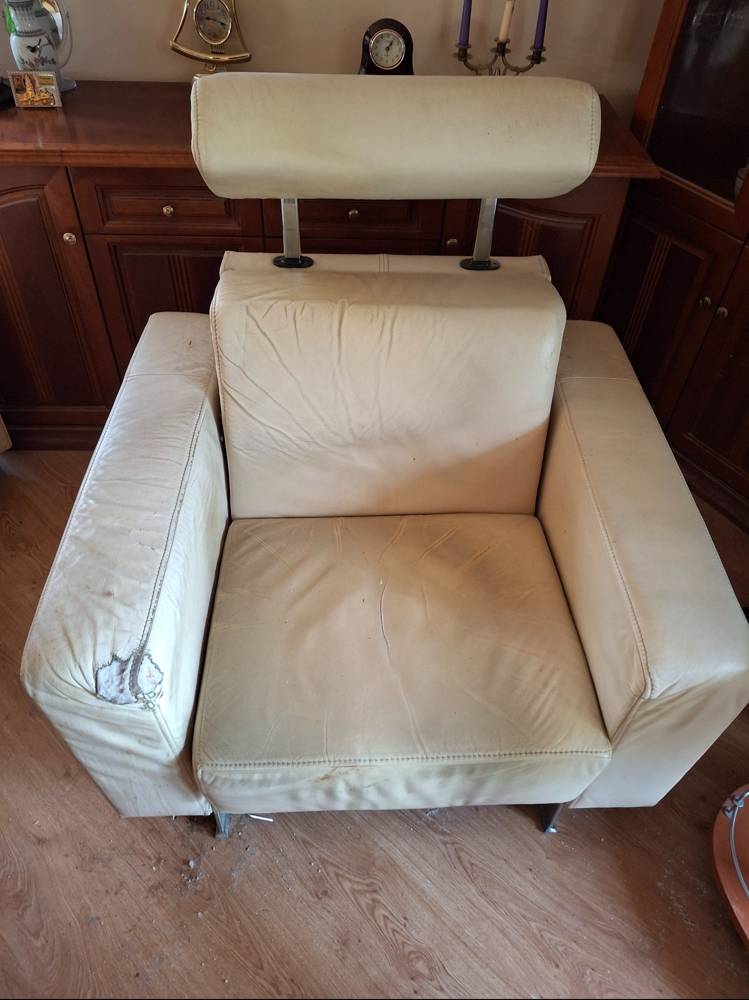 Beżowy fotel skórzany z widocznymi uszkodzeniami tapicerki na boku, stojący na drewnianej podłodze, z meblami w tle. Widoczne ślady użytkowania i zniszczenia.