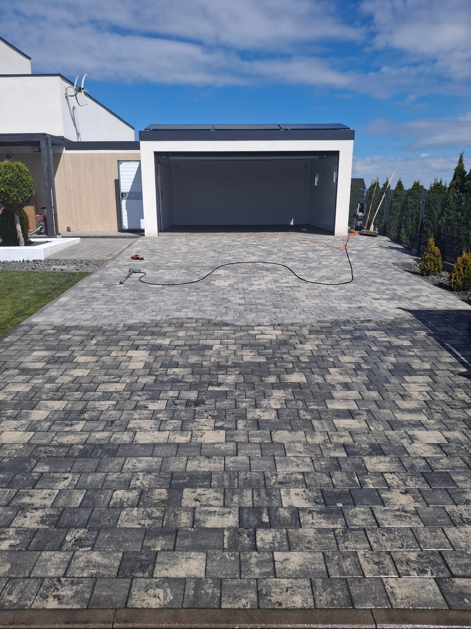 Czyszczenie kostki brukowej myjką ciśnieniową na podjeździe przed garażem. Widoczny efekt czyszczenia, ciemniejsza i jaśniejsza kostka. Kabel myjki na kostce.